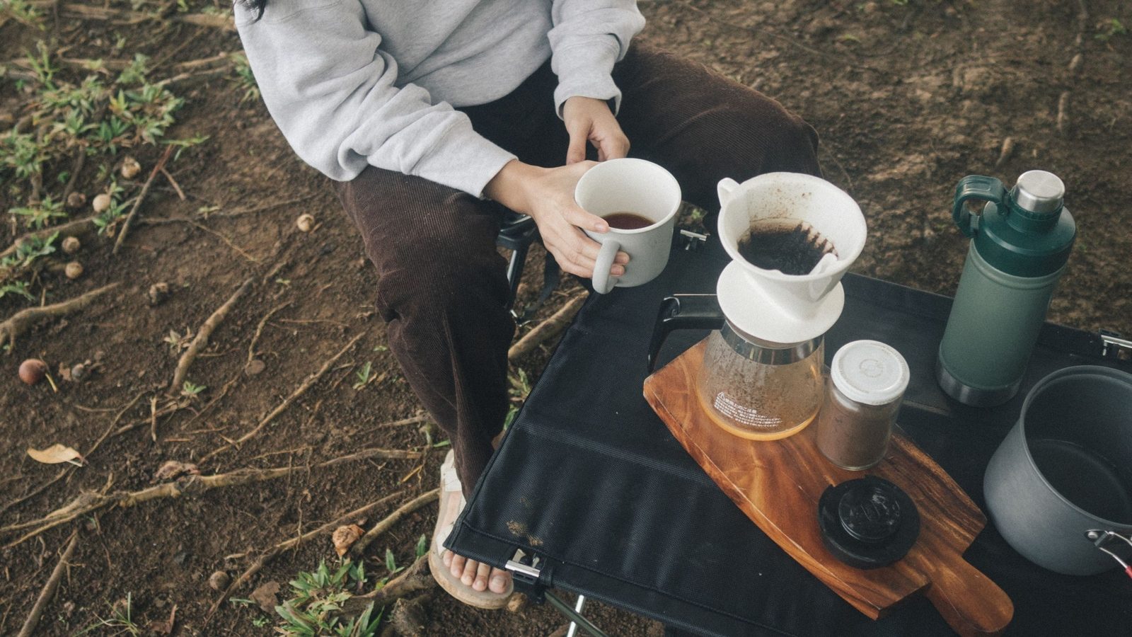 Frisch aufgebrühter Kaffee per Handfilter beim Camping – Person mit Kaffeetasse, Pour-Over-Dripper auf Glaskanne, Thermosflasche und Campingkocher auf einem Holzbrett im Freien.